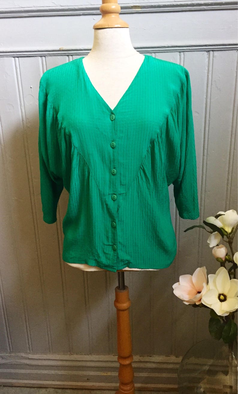 Puede incluir: Una blusa verde con botones, cuello en V y mangas 3/4. La blusa tiene un aspecto ligeramente fruncido y est&aacute; hecha de una tela sedosa.