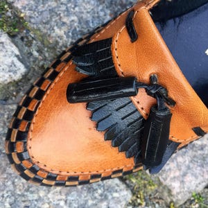 May include: Close-up of a brown leather loafer with black tassel detailing and a black and white checkered trim. The shoe has a size 4 label and says "Made in W-Germany".