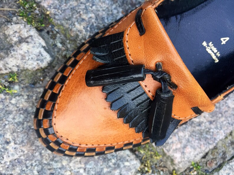 May include: Close-up of a brown leather loafer with black tassel detailing and a black and white checkered trim. The shoe has a size 4 label and says "Made in W-Germany".
