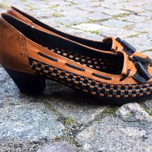 May include: Brown leather heeled shoes with black woven detailing and a tassel accent.