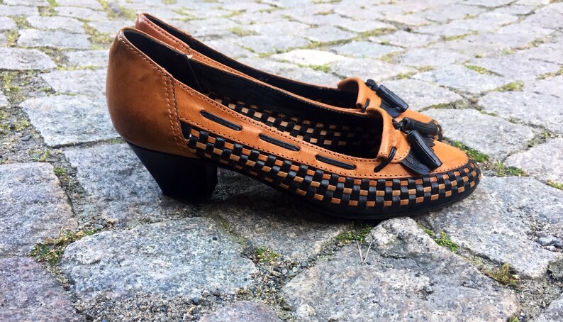 May include: Brown leather heeled shoes with black woven detailing and a tassel accent.