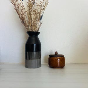 May include: A black and gray vase with dried flowers and a small, round, brown jar with a lid. The vase has a striped design on the lower half. The jar has a wooden knob on top.