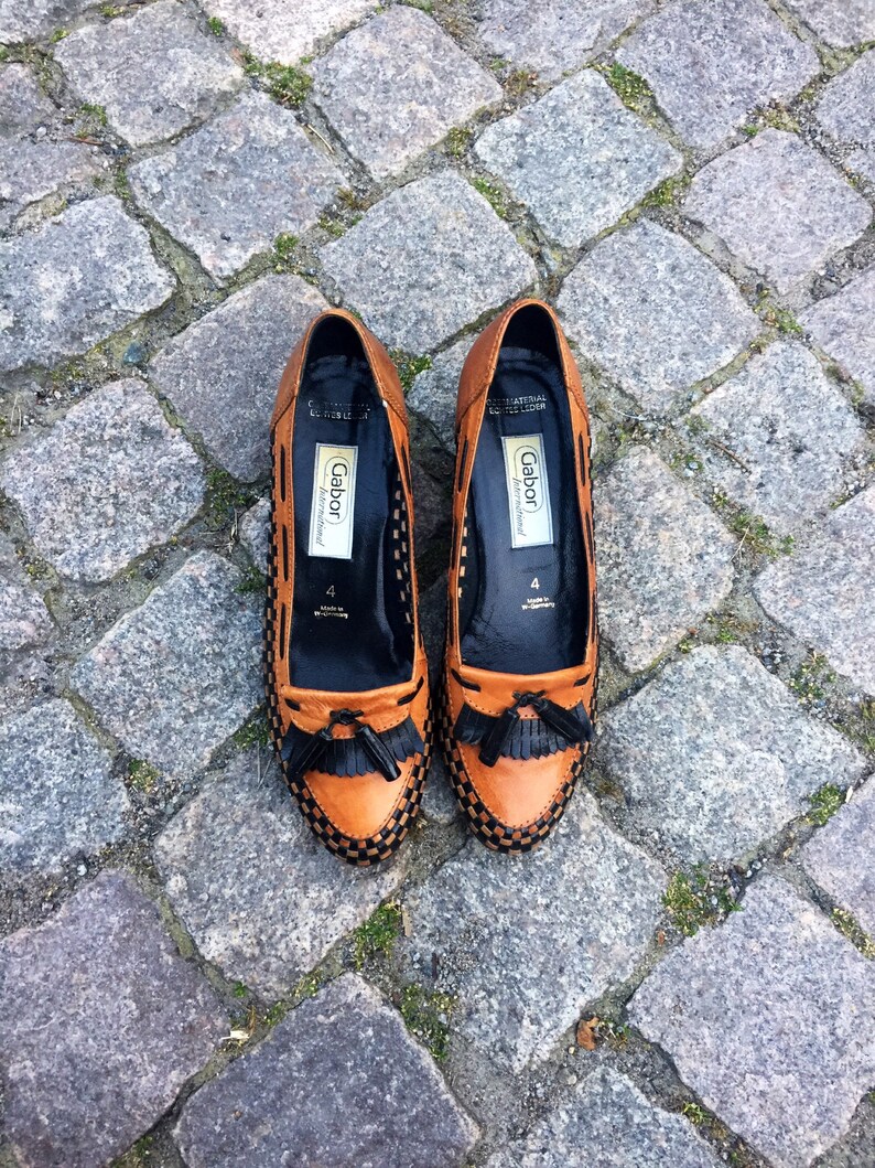 May include: A pair of brown leather loafers with black trim and tassels. The loafers have a woven pattern and the brand name "Gabor" is printed on the inside.