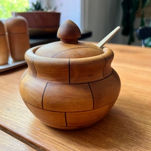 May include: A wooden honey pot with a lid and a small wooden spoon. The pot is brown and has a checkered pattern.
