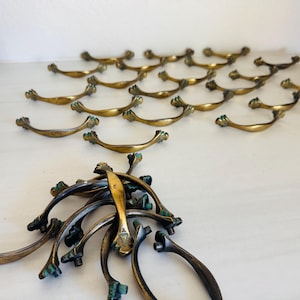May include: A collection of antique brass drawer pulls with a curved design. The pulls have a golden finish with green accents and are arranged on a white surface. Some pulls are grouped together, while others are neatly arranged.