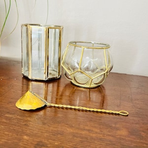 May include: Two gold-rimmed glass candle holders and a gold-colored metal stirring spoon on a wooden surface.