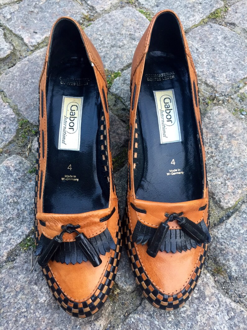 May include: A pair of brown leather loafers with black tassel accents. The shoes have a checkered pattern and are labeled "Gabor International" with the size "4" and "Made in W-Germany".