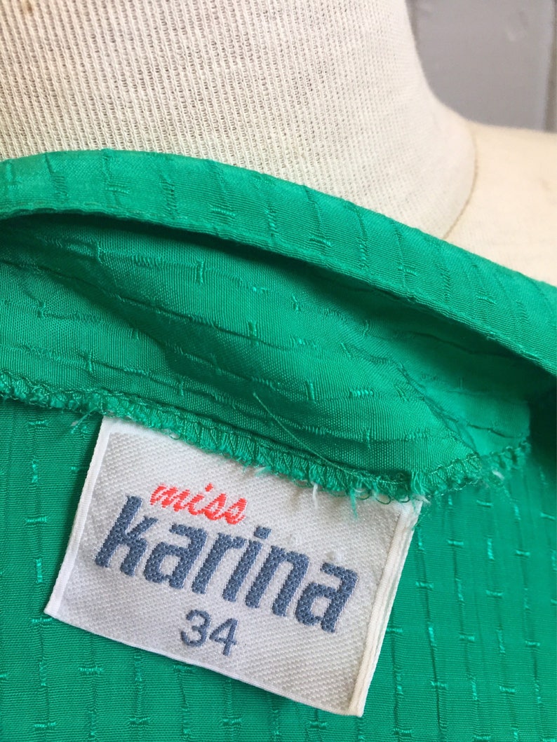 Puede incluir: Una tela verde texturizada con una etiqueta blanca que dice "miss Karina" y "34" en letras azules.