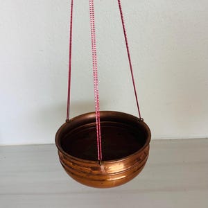 May include: A round copper bowl with a polished metallic finish and horizontal ridges along the exterior. The bowl is suspended by three thin red and white patterned cords attached to the rim, allowing it to hang in a balanced position.