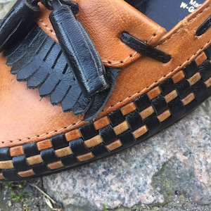 May include: Brown leather loafer with black woven trim and a black tassel. The loafer has a stitched seam around the edge.