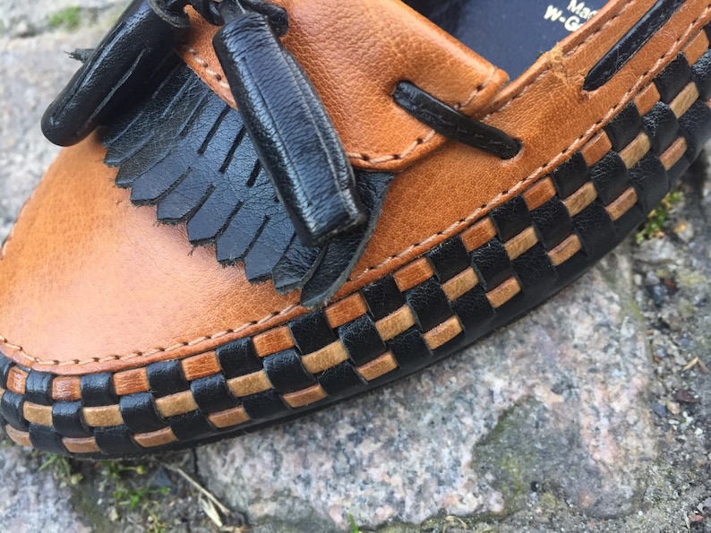 May include: Brown leather loafer with black woven trim and a black tassel. The loafer has a stitched seam around the edge.