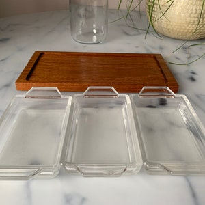 K&ouml;nnte beinhalten: Drei rechteckige Glasschalen mit abgerundeten Kanten und Griffen auf einem Holztablett. Die Schalen sind leer und bereit, mit Snacks oder Vorspeisen gef&uuml;llt zu werden.