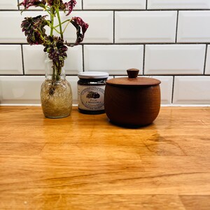 May include: A clear glass vase with a plant with dark red and green leaves, a jar of preserves, and a small wooden container with a lid sit on a wooden surface. The background is a white tiled wall.