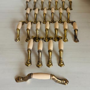 May include: A collection of cabinet pulls with a cream-colored center and brass-toned metal accents. The pulls are arranged on a light-colored surface, showcasing their design and quantity. The hardware is likely for furniture restoration or new construction.