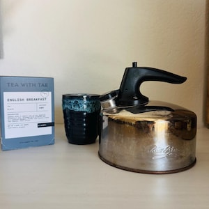 May include: A stainless steel kettle with a black handle and spout, a blue and black ceramic mug, and a box of "TEA WITH TAE" English Breakfast tea. The kettle has a vintage look.