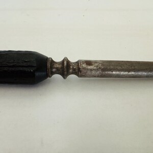 May include: Close-up of a vintage metal tool with a dark, textured handle. The handle leads to a decorative metal section, followed by a long, slender, and aged metal shaft. The tool appears to be well-worn.