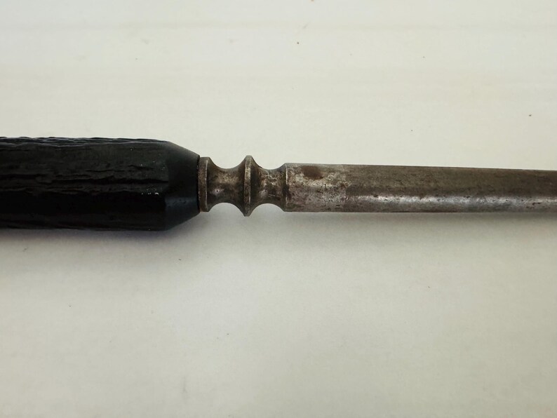 May include: Close-up of a vintage metal tool with a dark, textured handle. The handle leads to a decorative metal section, followed by a long, slender, and aged metal shaft. The tool appears to be well-worn.