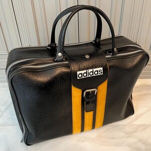 Op de afbeelding: Zwarte Adidas sporttas met een gele streep en het Adidas-logo. De tas heeft een handvat aan de bovenkant en een ritssluiting. De tas is gemaakt van een leerachtig materiaal.