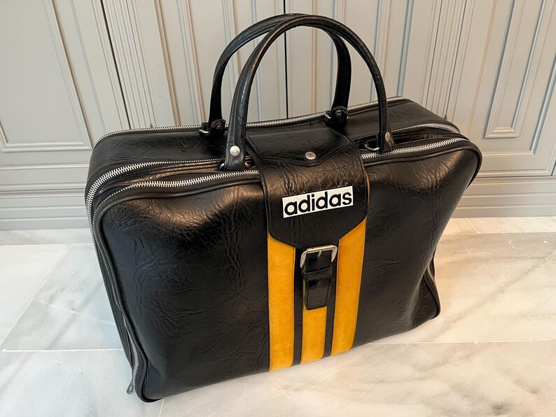 Op de afbeelding: Zwarte Adidas sporttas met een gele streep en het Adidas-logo. De tas heeft een handvat aan de bovenkant en een ritssluiting. De tas is gemaakt van een leerachtig materiaal.