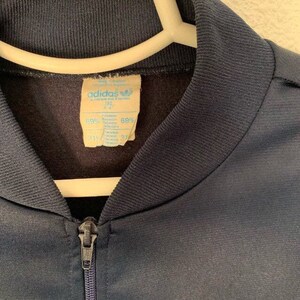 May include: A navy blue Adidas tracksuit jacket with a light blue stripe across the bottom. The jacket has a zipper closure and a label that reads "adidas" and "69% Polyester 31% Cotton".
