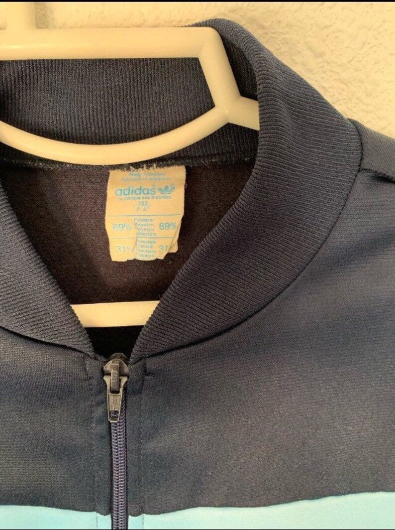 May include: A navy blue Adidas tracksuit jacket with a light blue stripe across the bottom. The jacket has a zipper closure and a label that reads "adidas" and "69% Polyester 31% Cotton".