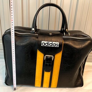 Op de afbeelding: Zwarte sporttas met gele strepen en het Adidas-logo. De tas heeft een handvat aan de bovenkant en een voorvak met een gesp. De tas is ongeveer 46 cm lang.