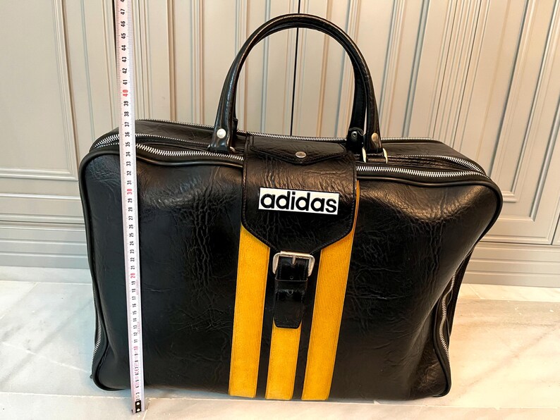 Op de afbeelding: Zwarte sporttas met gele strepen en het Adidas-logo. De tas heeft een handvat aan de bovenkant en een voorvak met een gesp. De tas is ongeveer 46 cm lang.