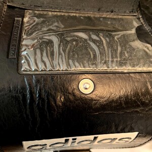 Op de afbeelding: Close-up van een zwarte Adidas-tas. De tas heeft een doorzichtige plastic zak, een zilveren drukknoopsluiting en het Adidas-logo in het wit. De woorden "MADE IN FRANCE" zijn zichtbaar aan de zijkant van de tas.