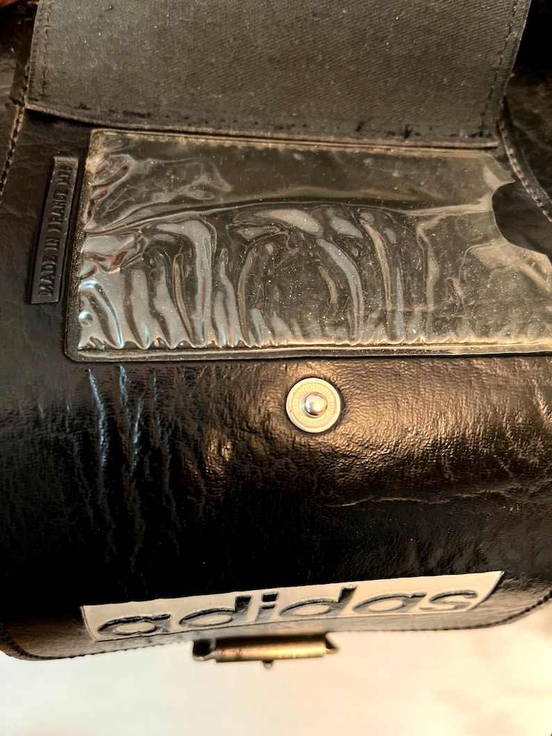 Op de afbeelding: Close-up van een zwarte Adidas-tas. De tas heeft een doorzichtige plastic zak, een zilveren drukknoopsluiting en het Adidas-logo in het wit. De woorden "MADE IN FRANCE" zijn zichtbaar aan de zijkant van de tas.