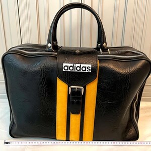 Op de afbeelding: Zwarte duffeltas met een gele streep en drie verticale gele strepen. De tas heeft een handvat aan de bovenkant, een ritssluiting en het woord "adidas" in het wit. De tas is ongeveer 61 cm breed.