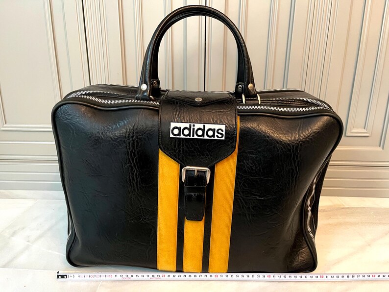 Op de afbeelding: Zwarte duffeltas met een gele streep en drie verticale gele strepen. De tas heeft een handvat aan de bovenkant, een ritssluiting en het woord "adidas" in het wit. De tas is ongeveer 61 cm breed.