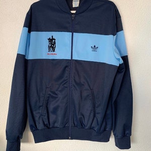 May include: A navy blue Adidas track jacket with a light blue stripe and the text "PHILADELPHIA" in red. The Adidas logo is on the right side of the jacket.