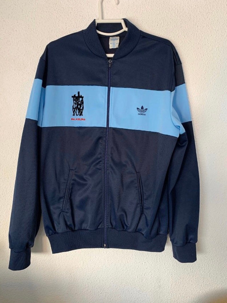 May include: A navy blue Adidas track jacket with a light blue stripe and the text "PHILADELPHIA" in red. The Adidas logo is on the right side of the jacket.