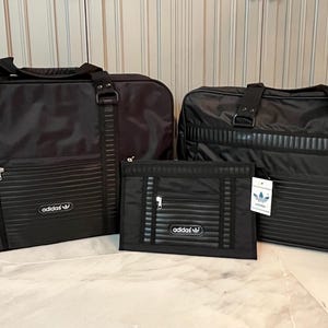 May include: Three-piece black luggage set. The set includes two large travel bags and a smaller document case. Each piece features a ribbed design, a zipper closure, and the Adidas logo. The bags have handles and straps.