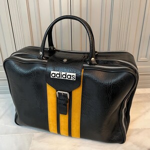 Op de afbeelding: Zwarte Adidas sporttas met een gele streep en het Adidas logo. De tas heeft een handvat aan de bovenkant, een ritssluiting en een gespsluiting. De tas is gemaakt van een textuur.