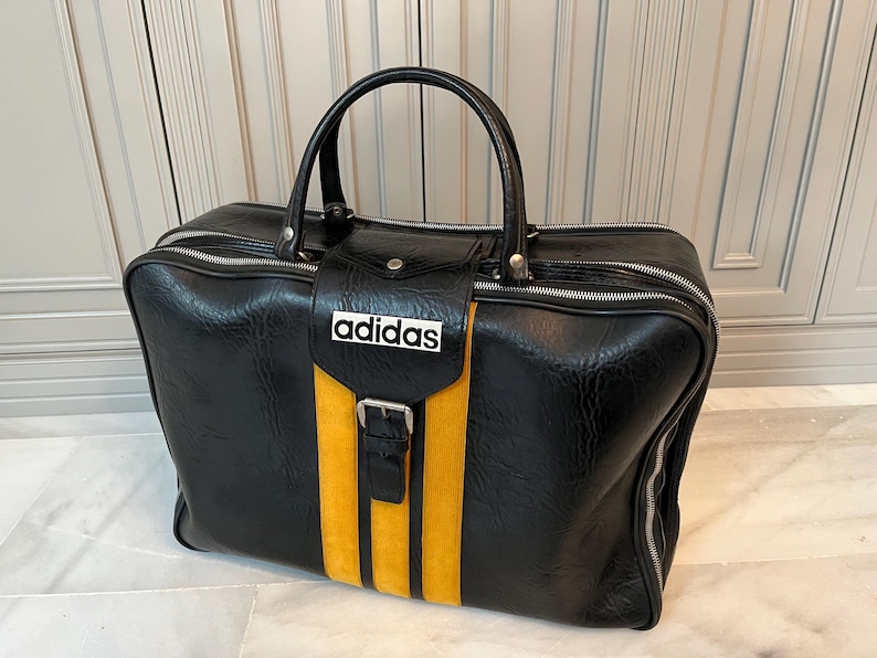 Op de afbeelding: Zwarte Adidas sporttas met een gele streep en het Adidas logo. De tas heeft een handvat aan de bovenkant, een ritssluiting en een gespsluiting. De tas is gemaakt van een textuur.