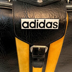 Op de afbeelding: Zwarte Adidas sporttas met gele strepen en het Adidas logo. De tas heeft een handvat aan de bovenkant, een klepsluiting met een gesp en een ritssluiting. De tas is gemaakt van een textuur.