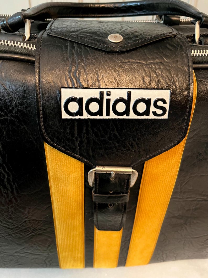 Op de afbeelding: Zwarte Adidas sporttas met gele strepen en het Adidas logo. De tas heeft een handvat aan de bovenkant, een klepsluiting met een gesp en een ritssluiting. De tas is gemaakt van een textuur.