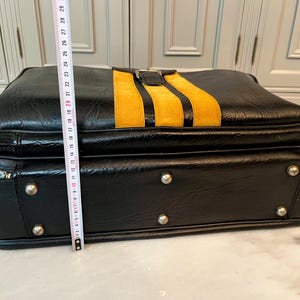 Op de afbeelding: Zwarte vintage aktetas met een gele corduroy streep. De aktetas heeft zilverkleurige hardware, inclusief ritsen en klinknagels. De hoogte van de koffer is ongeveer 25 cm.