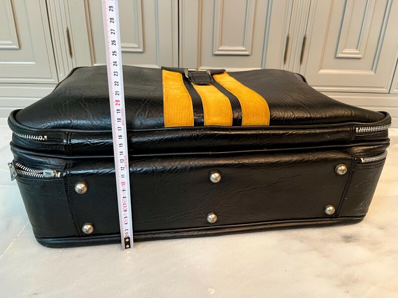 Op de afbeelding: Zwarte vintage aktetas met een gele corduroy streep. De aktetas heeft zilverkleurige hardware, inclusief ritsen en klinknagels. De hoogte van de koffer is ongeveer 25 cm.