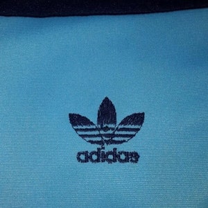 May include: A blue athletic shirt with a navy blue Adidas logo featuring the company's signature three-leaf clover design and the word "adidas" underneath.