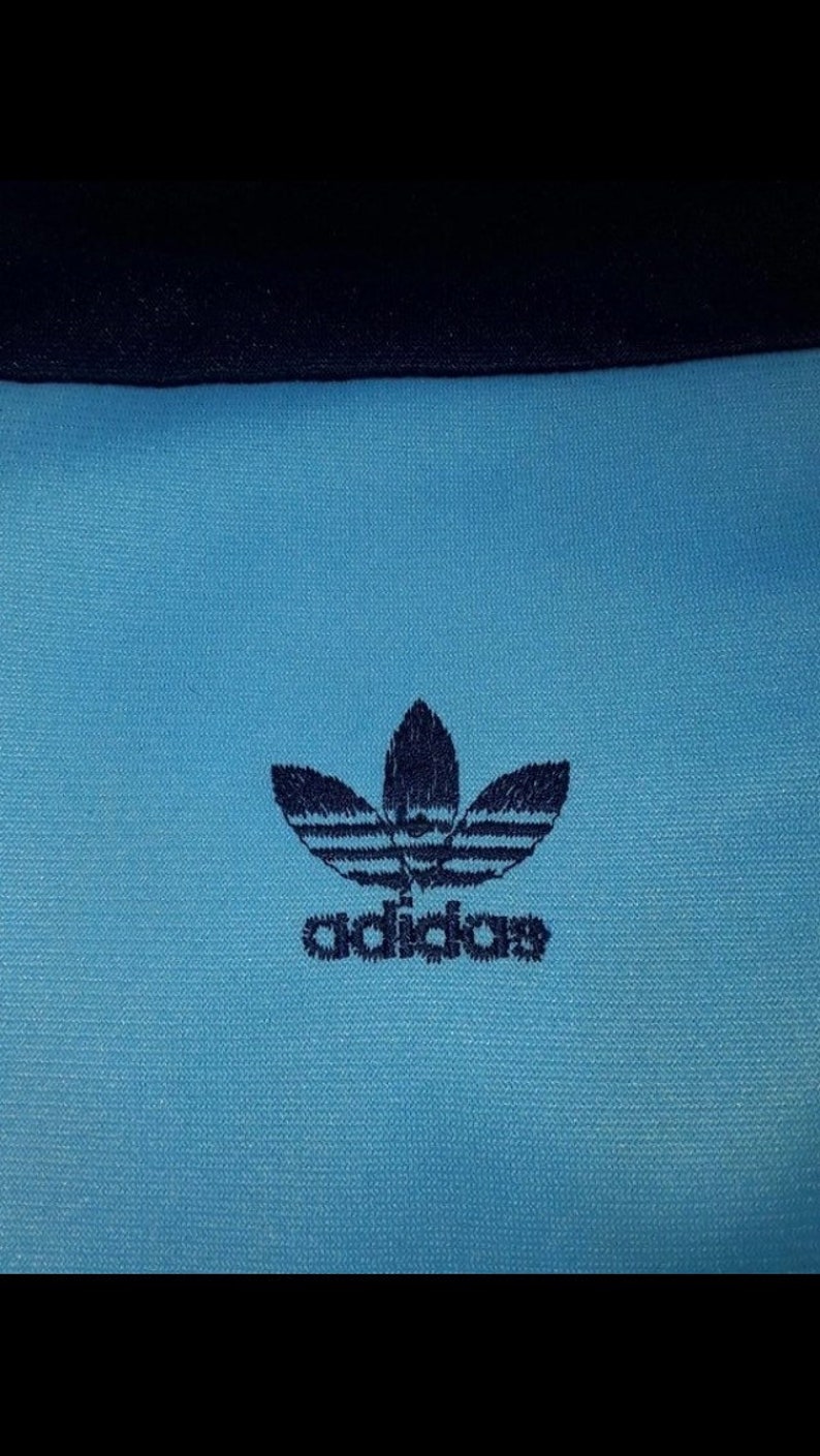 May include: A blue athletic shirt with a navy blue Adidas logo featuring the company's signature three-leaf clover design and the word "adidas" underneath.