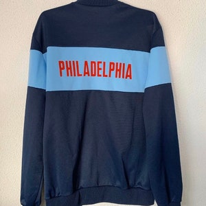 May include: A navy blue zippered jacket with a light blue stripe across the back. The stripe has the word "PHILADELPHIA" printed in red.