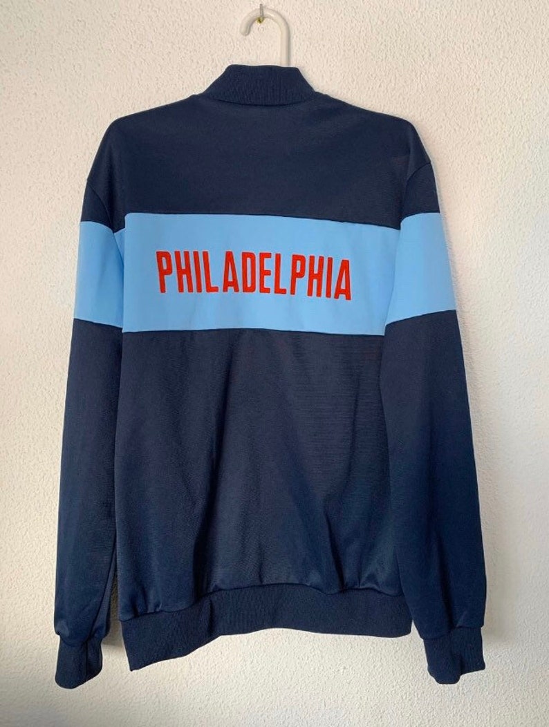 May include: A navy blue zippered jacket with a light blue stripe across the back. The stripe has the word "PHILADELPHIA" printed in red.