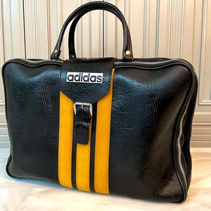 Op de afbeelding: Zwarte Adidas reistas met een gele streep in het midden. De tas heeft een handvat aan de bovenkant en een zilveren ritssluiting. Het Adidas-logo staat op de voorkant. De tas is gemaakt van een textuur.
