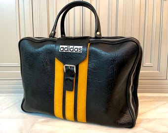 Puede incluir: Bolso de viaje Adidas negro con una franja amarilla en el centro. El bolso tiene un asa superior y una cremallera plateada. El logotipo de Adidas se muestra en la parte delantera. El bolso está hecho de un material texturizado.