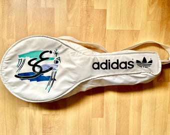 May include: Off-white tennis racket bag with a shoulder strap. The bag features the Adidas logo and a colorful graphic design on the front. The graphic includes teal, blue, and black elements.