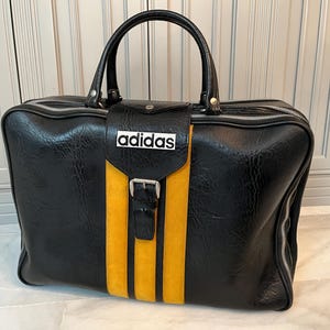 Op de afbeelding: Zwarte Adidas sporttas met een gele streep in het midden en het Adidas-logo. De tas heeft een handvat aan de bovenkant, een gespsluiting en een ritssluiting. Gemaakt van een textuur materiaal. Afmetingen: ca. 50 cm x 30 cm x 25 cm.