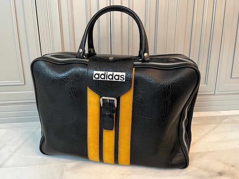 Op de afbeelding: Zwarte Adidas sporttas met een gele streep in het midden en het Adidas-logo. De tas heeft een handvat aan de bovenkant, een gespsluiting en een ritssluiting. Gemaakt van een textuur materiaal. Afmetingen: ca. 50 cm x 30 cm x 25 cm.