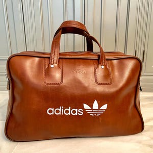 May include: A brown leather Adidas duffel bag with a white Adidas logo and text. The bag has two handles and a zipper closure. The bag is approximately 20 inches long, 10 inches high, and 10 inches wide.